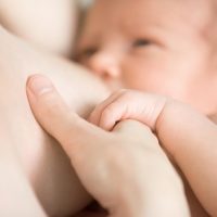 Close-up portrait of sweet newborn baby breastfeeding. Adorable new born caucaian kid holding moms finger with his cute little palm while breast feeding. Focus on hands. Young mother nursing baby